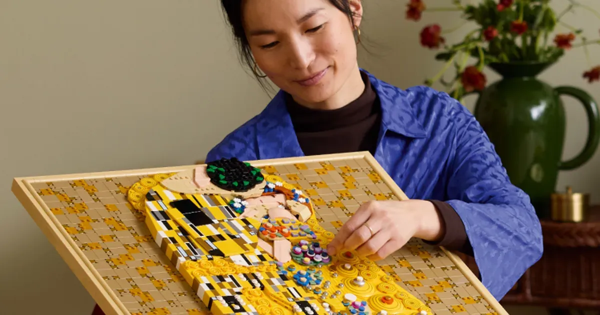 Kvinna i blå jacka tittar på en LEGO-konstbyggsats föreställande Klimts målning "Kyssen" med gyllene och svarta detaljer och färgglada pärlor, framför en vas me